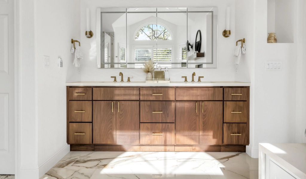 A bright, modern bathroom featuring a wooden vanity with gold-toned fixtures, a large mirror, and marble flooring.