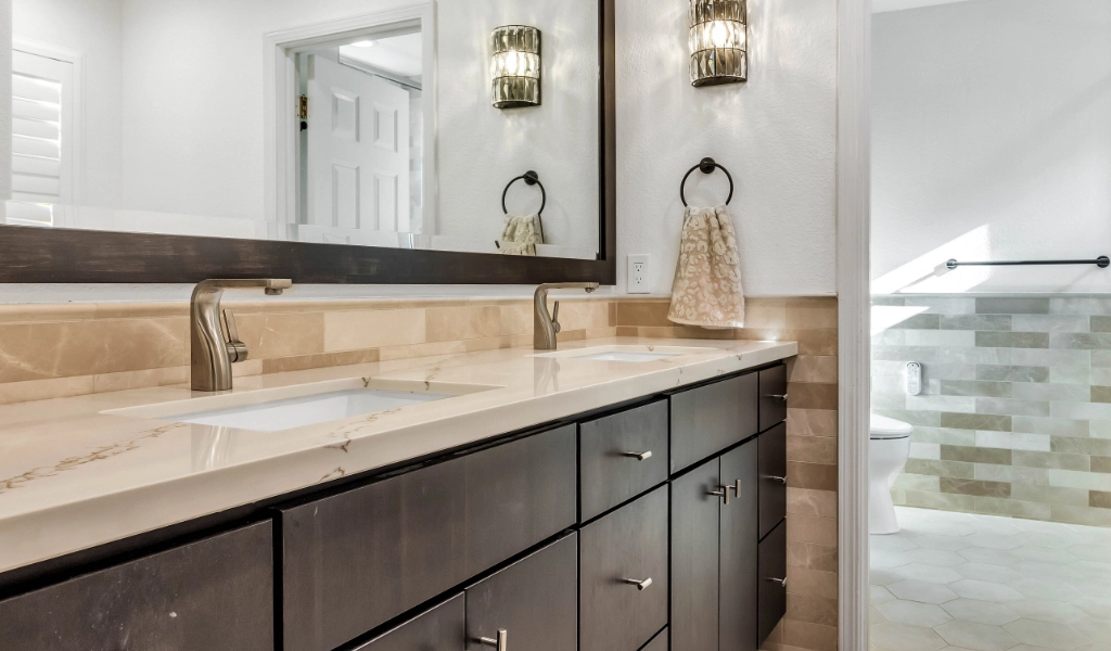 A modern bathroom with dual sinks, dark wood vanity, and beige countertops, featuring wall-mounted lamps and a glass-enclosed shower in the background.