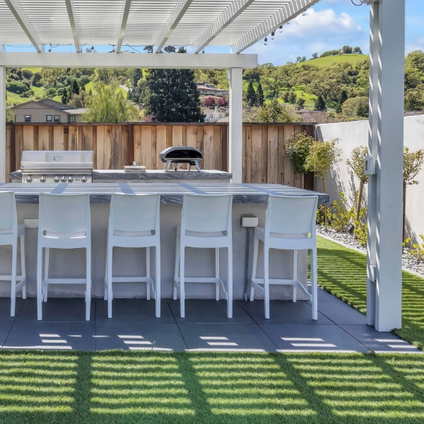 an outdoor living kitchen space with a white pergola, dining table, and seating, surrounded by lush greenery.