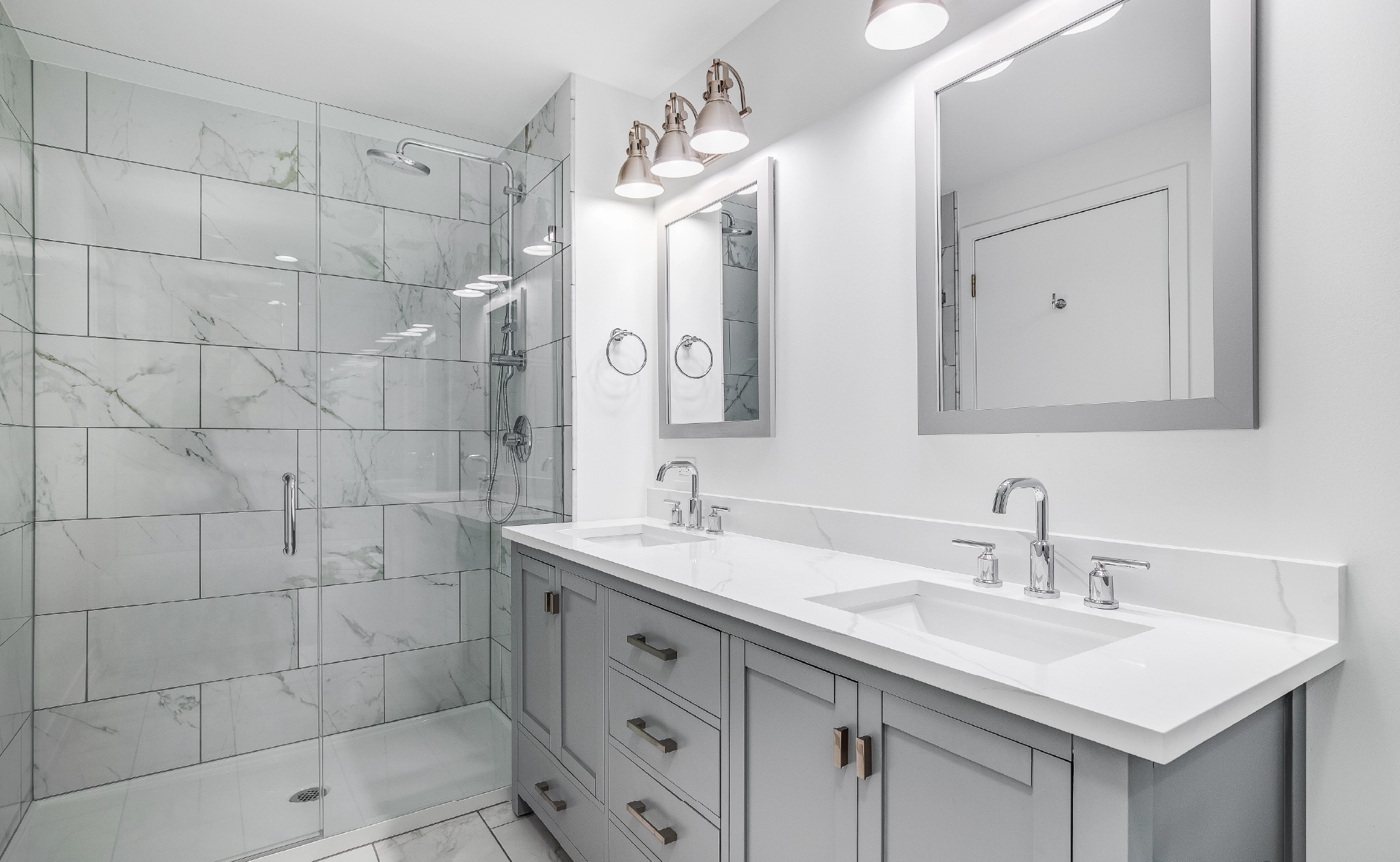 An elegant, remodeled bathroom with a grey vanity and bronze hardware. The shower has a large shower head and marble tiles and glass wall line the sides.