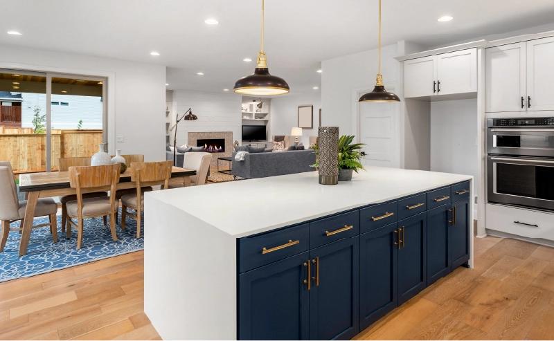 Modern kitchen layout with a white island featuring navy blue cabinets, pendant lights above, adjacent dining area, and view into a living room with fireplace.