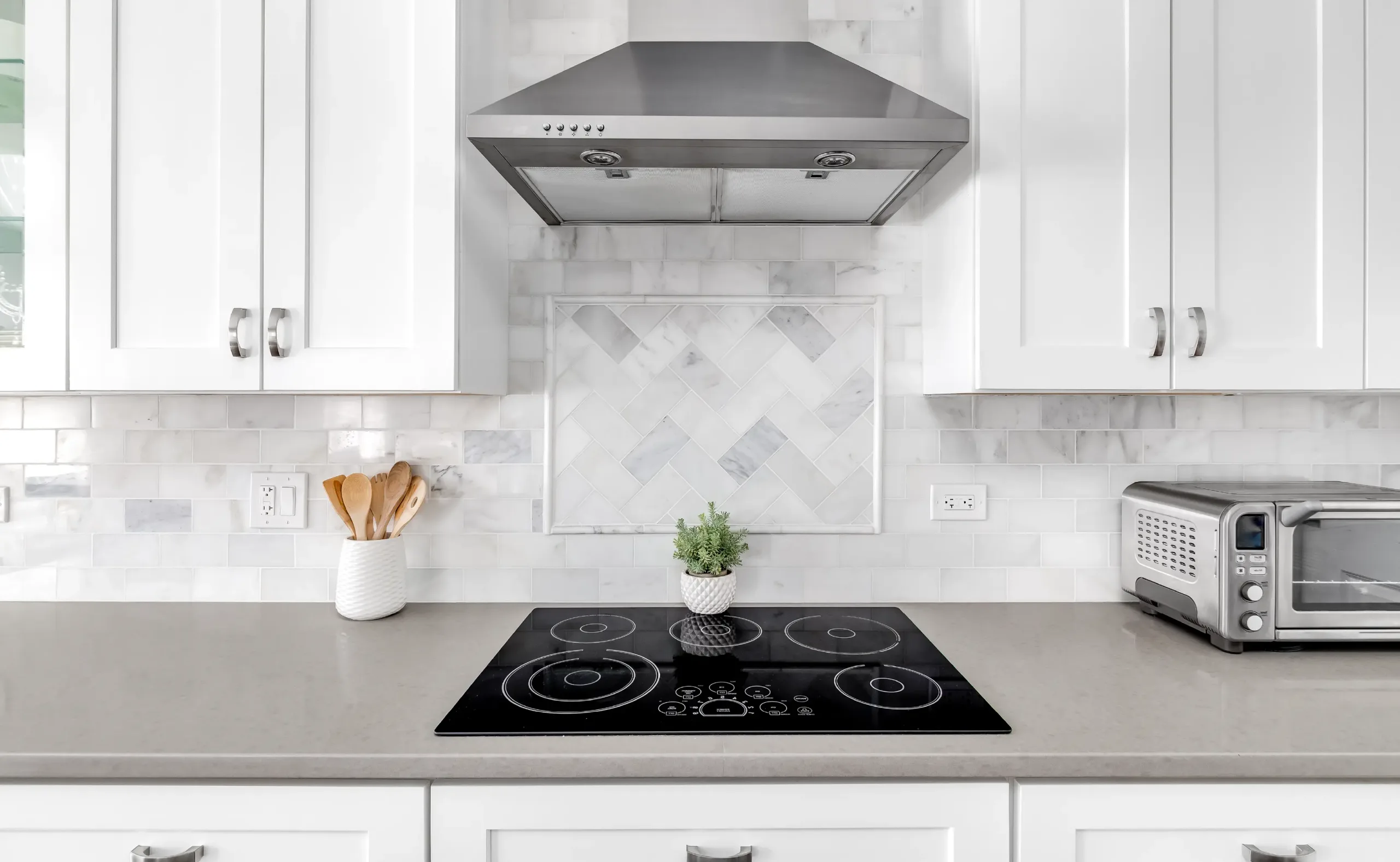 Modern kitchen with white cabinets, stainless steel range hood, black glass electric cooktop, marble tile backsplash, and countertop appliances including a toaster oven and utensil holder.