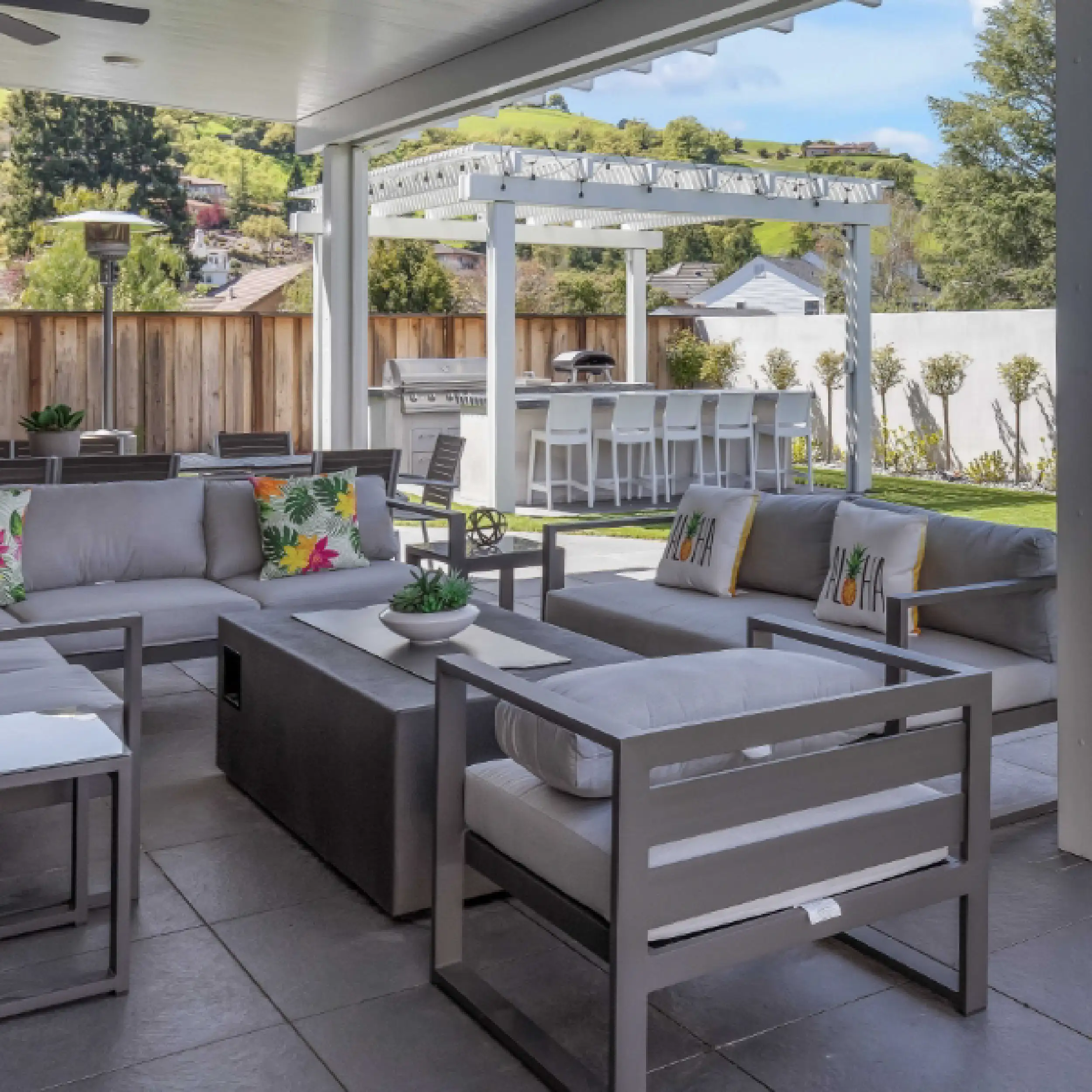 Modern outdoor patio with gray furniture, tropical accent pillows, a covered seating area, a pergola over an Outdoor Kitchen and dining space, all set in a fenced backyard.