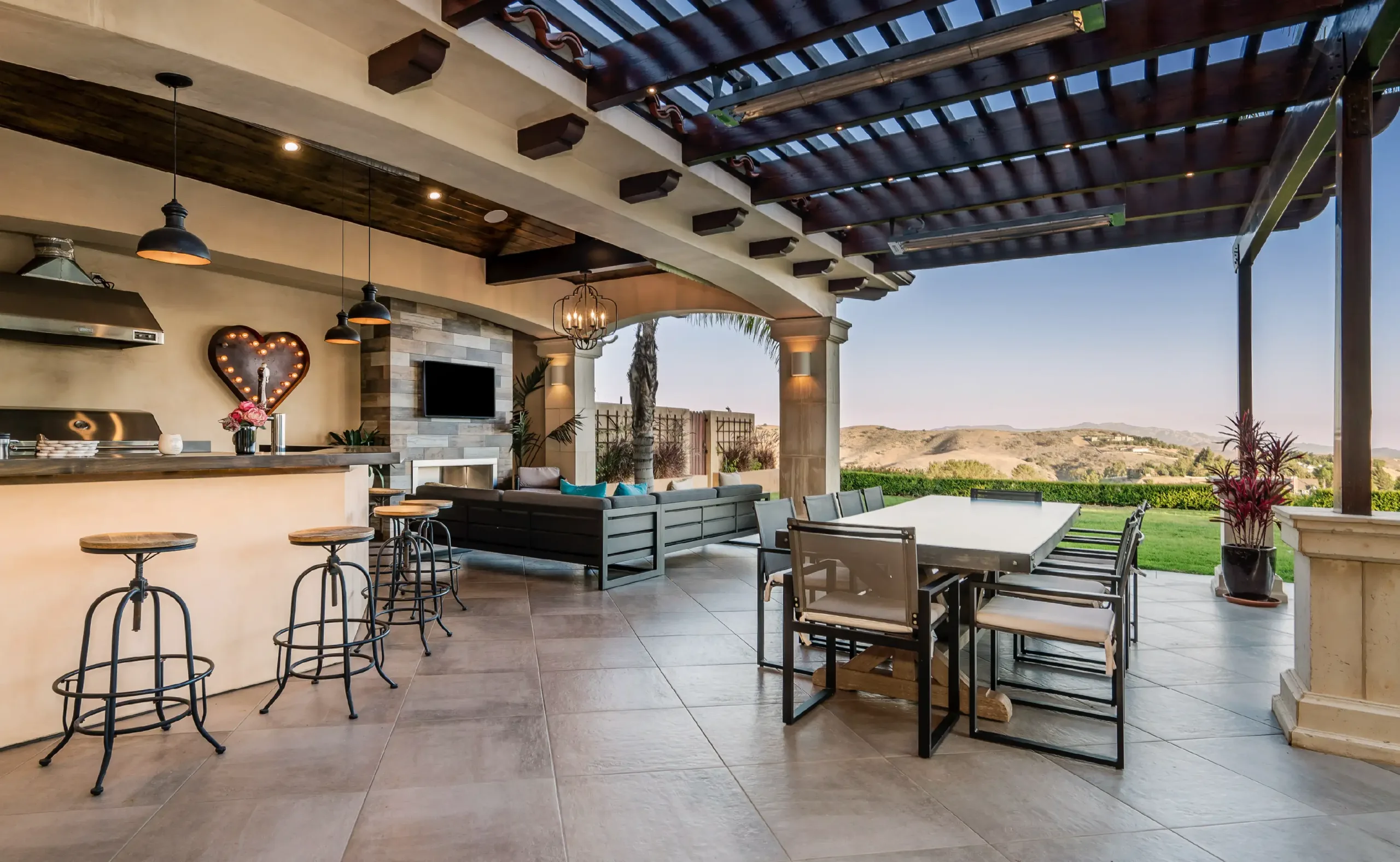 Spacious outdoor patio with an Outdoor Kitchen, bar stools, dining table, and lounge area under a pergola, overlooking hills and greenery.
