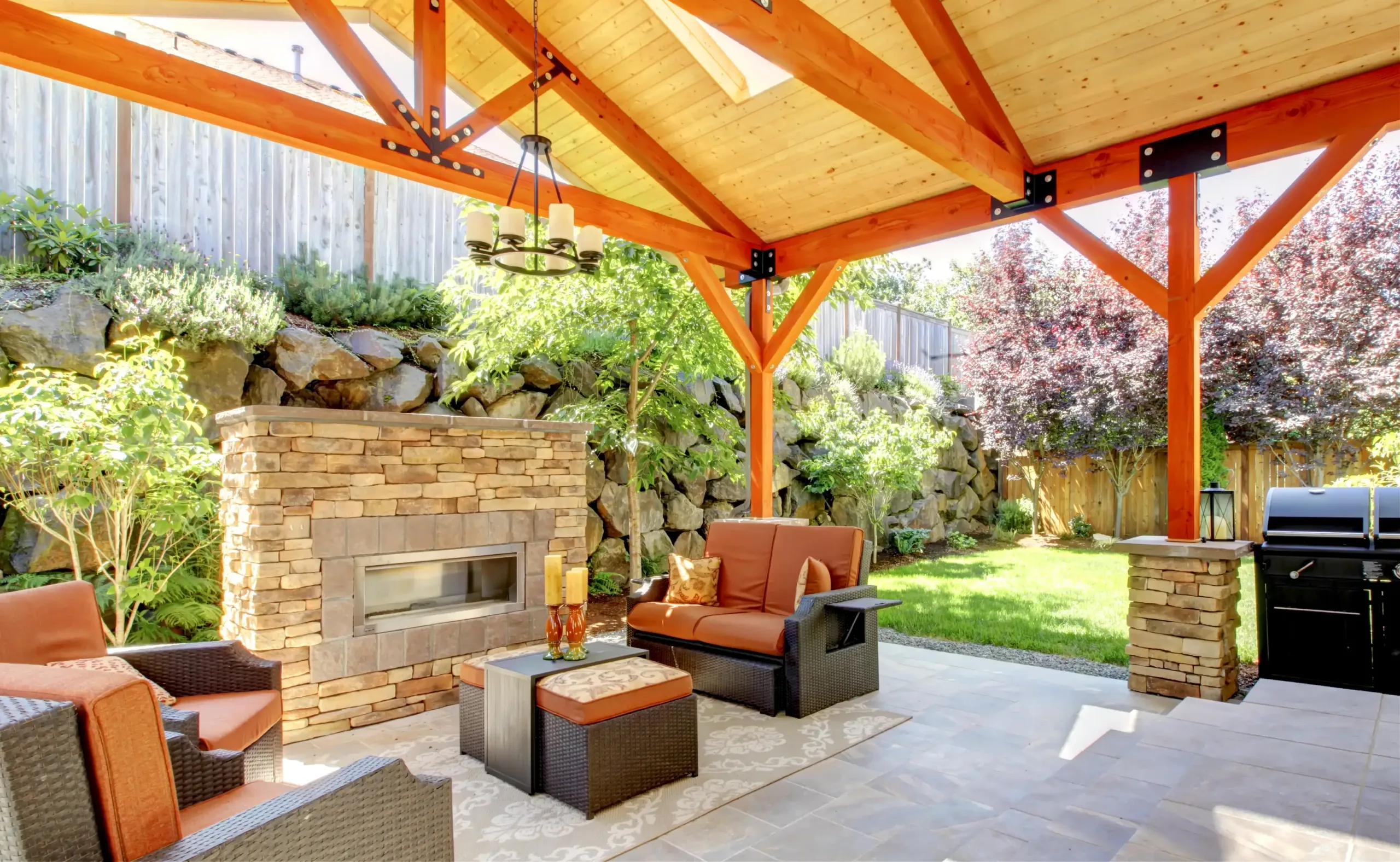 Covered outdoor patio with wooden beams, stone fireplace, and wicker furniture with orange cushions. Enjoy a rug, grill, and beautifully landscaped backyard views—perfect for relaxing or entertaining outdoors.