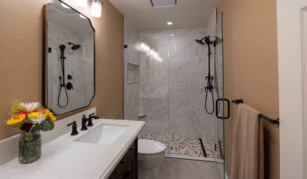 Modern bathroom with a white vanity, dual sinks, a glass-enclosed shower with marble walls, dark fixtures, beige walls, and a towel on a rack. Thoughtfully designed for a Kids' Bathroom Remodel, it features practical touches like easy-to-reach counters and fun accents. A small flower vase adds charm to the counter.