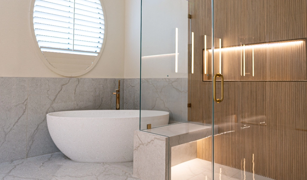 Modern Primary & Hall Bathroom Remodel featuring a freestanding white tub, marble floor and walls, wood-paneled shower with glass door, and a round window with shutters.
